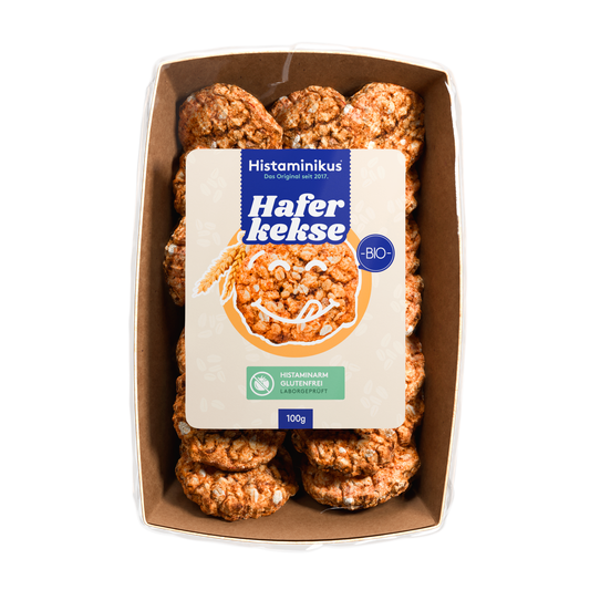 Rechteckige Kartonverpackung mit mehreren runden Haferkeksen. Auf der Vorderseite ein weiß-blaues Etikett mit der Aufschrift „Haferkekse – Bio“. Darunter Symbole und Hinweise: „Histaminarm“, „Glutenfrei“, „Laborgeprüft“. Verpackungsgewicht: 100 g. Design mit gezeichnetem Smiley auf einem Keks und einer Ähre als grafisches Element. 