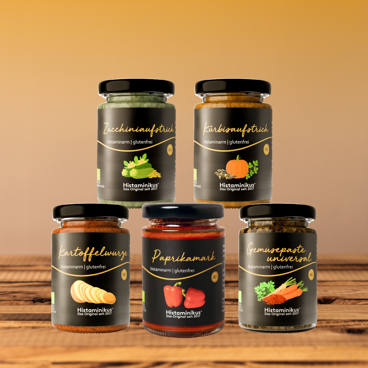 Fünf herzhafte Produkte von Histaminikus in Gläsern auf Holztisch: Zucchiniaufstrich, Kürbisaufstrich, Kartoffelwürze, Paprikamark und Gemüsepaste universal, vor warmem Farbverlauf-Hintergrund.