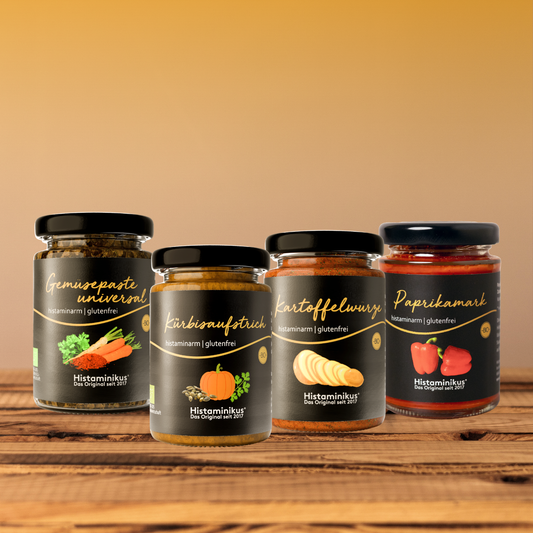 Vier Histaminikus-Gläser auf einem Holztisch vor warmem, unscharfem Hintergrund: Gemüsepaste universal, Kürbisaufstrich, Kartoffelwürze und Paprikamark. Alle Produkte sind frontal angeordnet und tragen schwarze Etiketten mit goldener Schrift sowie den Hinweis „histaminarm | glutenfrei“.
