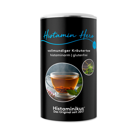 Schwarze Teedose mit der Aufschrift „Histamin Hero – vollmundiger Kräutertee“, darunter „histaminarm | glutenfrei“. In der Mitte ein rundes Foto mit dampfender Tasse Kräutertee auf einem Holzuntergrund, daneben zwei kleine Pflanzenabbildungen.