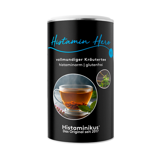 Schwarze Teedose mit der Aufschrift „Histamin Hero – vollmundiger Kräutertee“, darunter „histaminarm | glutenfrei“. In der Mitte ein rundes Foto mit dampfender Tasse Kräutertee auf einem Holzuntergrund, daneben zwei kleine Pflanzenabbildungen.