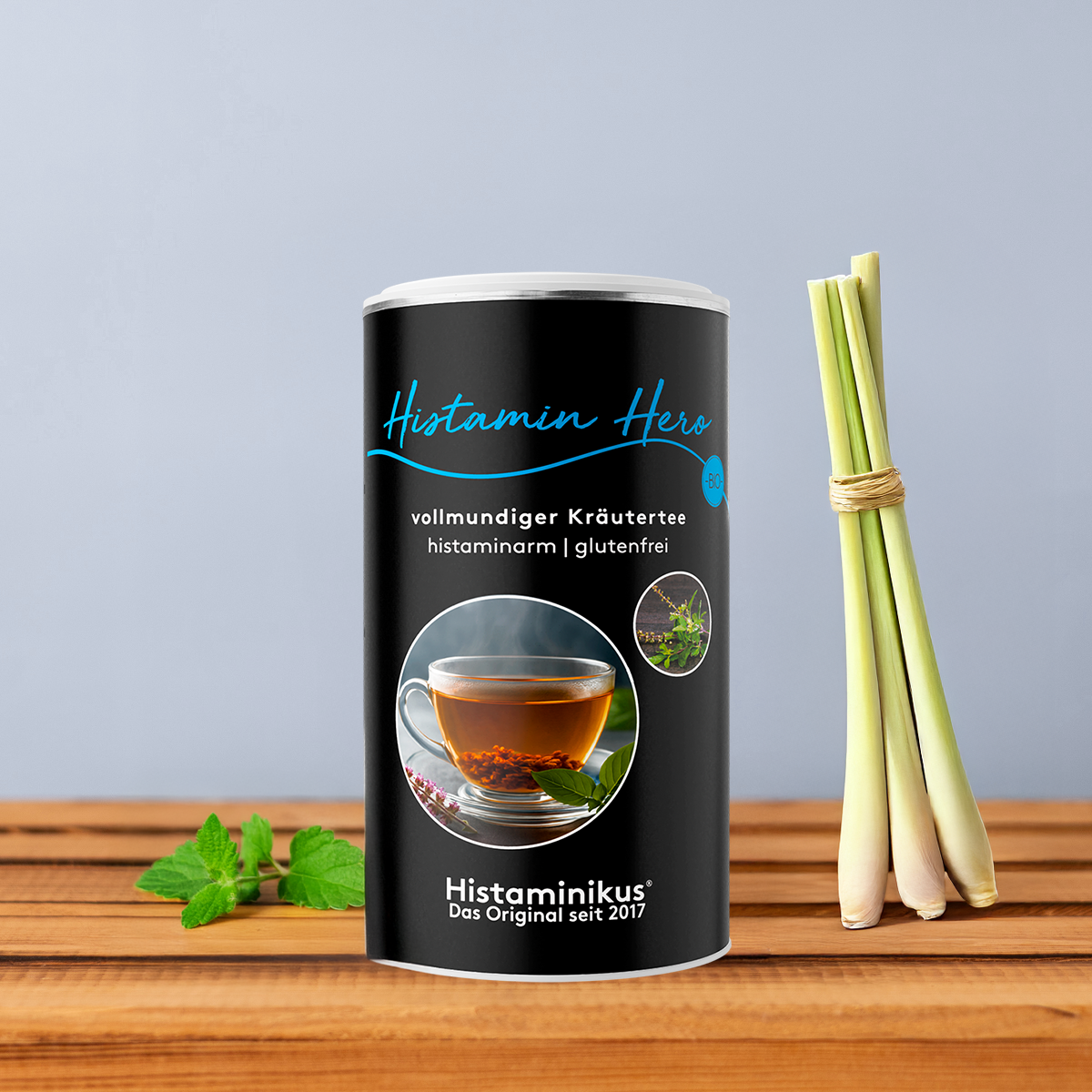 Schwarze Teedose „Histamin Hero“ mit blauer Schrift und Abbildung einer dampfenden Teetasse, dekoriert mit Zitronengras und Minze vor graublauem Hintergrund. 