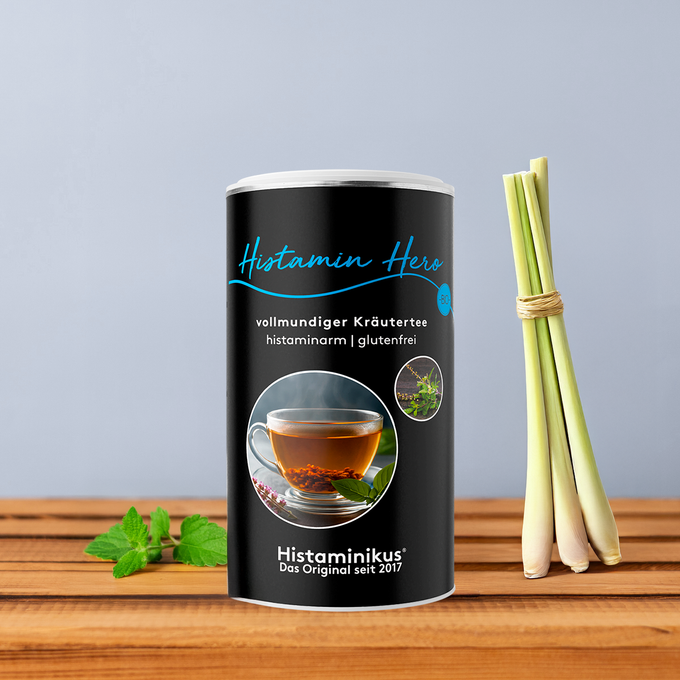 Schwarze Teedose „Histamin Hero“ mit blauer Schrift und Abbildung einer dampfenden Teetasse, dekoriert mit Zitronengras und Minze vor graublauem Hintergrund. 
