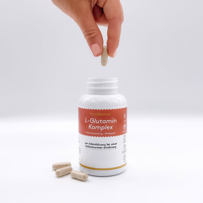 Eine Hand mit hellrosa lackierten Fingern hält eine Kapsel über eine geöffnete Dose mit der Aufschrift „HistaBalance® L-Glutamin Komplex“. Die Dose steht auf einer weißen Fläche, daneben liegen einige Kapseln. Das Etikett ist rot-weiß gestaltet und enthält Angaben wie „1 Monatspackung – 90 Kapseln“ und „zur Unterstützung bei einer histaminarmen Ernährung“. 