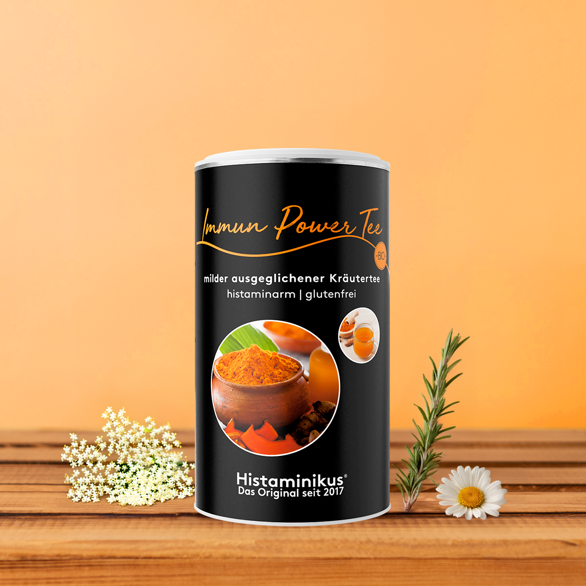 Zylinderdose „Immun Power Tee“ von Histaminikus auf einem Holzuntergrund vor orangem Hintergrund, flankiert von Holunderblüten, Rosmarinzweig und Kamillenblüte. Auf der Dose steht: „Immun Power Tee – milder ausgeglichener Kräutertee, histaminarm, glutenfrei“. Abgebildet sind Kurkumapulver und eine Tasse Tee. 