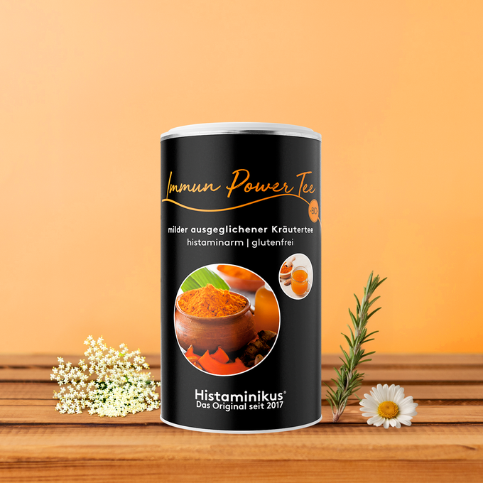 Zylinderdose „Immun Power Tee“ von Histaminikus auf einem Holzuntergrund vor orangem Hintergrund, flankiert von Holunderblüten, Rosmarinzweig und Kamillenblüte. Auf der Dose steht: „Immun Power Tee – milder ausgeglichener Kräutertee, histaminarm, glutenfrei“. Abgebildet sind Kurkumapulver und eine Tasse Tee. 