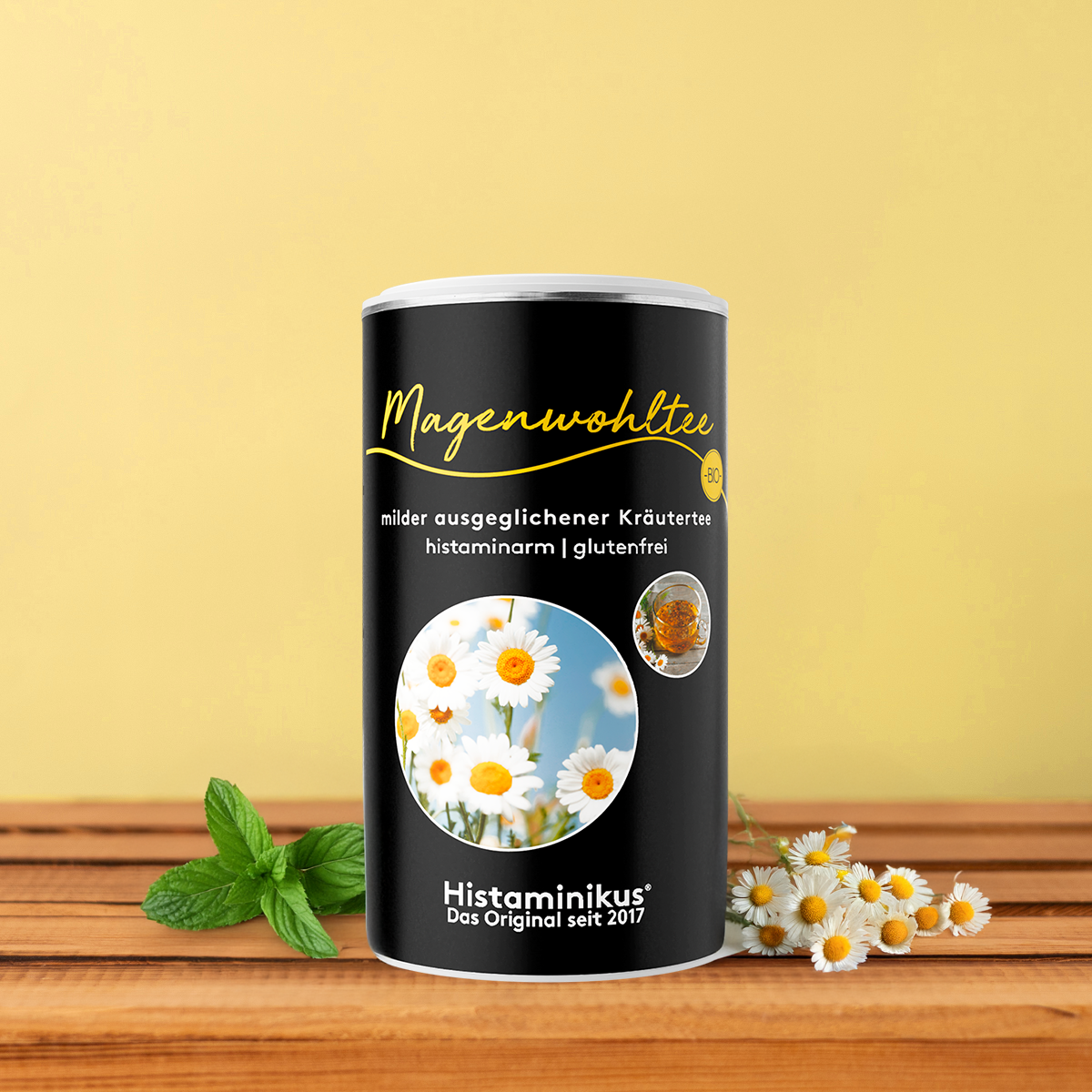 Schwarze Teedose „Magenwohltee“ mit gelber Schrift und Abbildung einer Teetasse, dekoriert mit Kamillenblüten und Minze vor gelbem Hintergrund. 