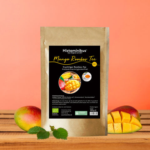 Standbeutel mit der Aufschrift „Histaminikus Mango Rooibos Tee – fruchtiger Rooibos-Tee, histaminarm, glutenfrei, BIO“. Auf dem Etikett sind eine aufgeschnittene Mango, Mangostücke und eine Tasse Tee abgebildet. Der Beutel steht auf einer Holzoberfläche vor einem orangefarbenen Hintergrund, links und rechts liegen ganze und aufgeschnittene Mangos sowie grüne Blätter.