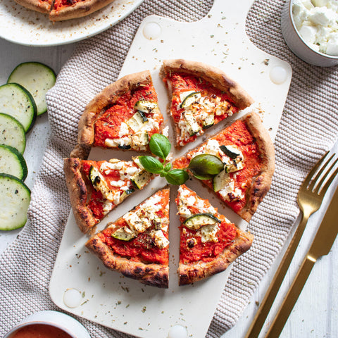 Produktbild der zubereiteten „Pizzaminikus“ Pizza mit knusprigem Rand, goldbraun gebacken. Belegt mit cremigem Hirtenkäse, Zucchinischeiben und frischem Basilikum auf fruchtiger Tomatensoße. Im Hintergrund eine Schale mit Feta, Zucchinischeiben und goldfarbenes Besteck auf heller Tischdecke.