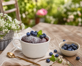 Schoko-Tassenkuchen in einer weißen Tasse, angerichtet auf einem rustikalen Holztisch im Garten. Der Kuchen ist mit Puderzucker bestäubt und mit Heidelbeeren sowie einem Minzblatt dekoriert. Daneben liegen einzelne Heidelbeeren, ein goldener Löffel und ein helles Stofftuch; im Hintergrund sind unscharfe Gartenpflanzen und Blumen zu sehen.