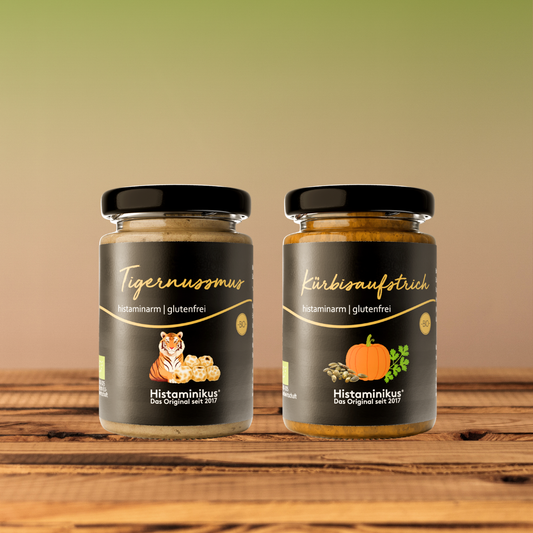 Zwei Histaminikus-Gläser auf einem Holztisch vor warmem, unscharfem Hintergrund: links Tigernussmus mit Tiger-Illustration, rechts Kürbisaufstrich mit Kürbis- und Kernmotiv. Beide Etiketten zeigen „histaminarm | glutenfrei“.