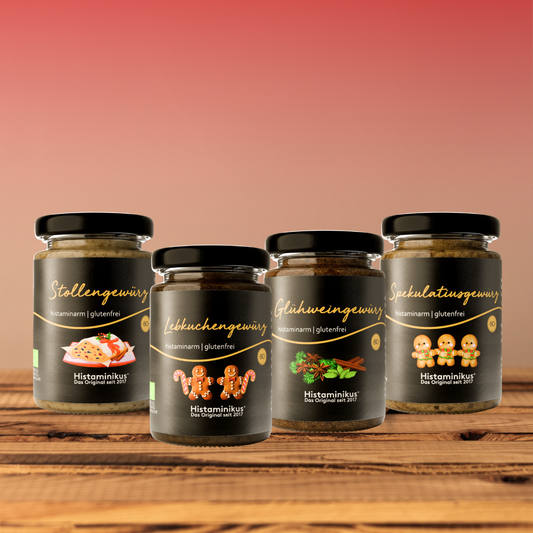 Vier histaminarme, glutenfreie Gewürzmischungen von Histaminikus in Glasgläsern: Stollengewürz, Lebkuchengewürz, Glühweingewürz und Spekulatiusgewürz, arrangiert auf einem Holztisch vor warmem Farbverlauf-Hintergrund.