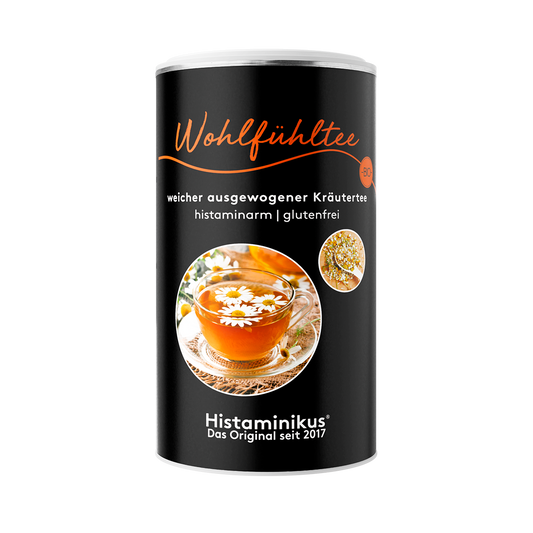 Schwarze Teedose mit der Aufschrift „Wohlfühltee – weicher ausgewogener Kräutertee – histaminarm | glutenfrei“. Auf dem Etikett ein großes rundes Bild mit einer Glastasse gefüllt mit Kräutertee, dekoriert mit Kamillenblüten, daneben ein kleineres Bild mit getrockneten Blüten. Unten das Logo „Histaminikus – Das Original seit 2017“.