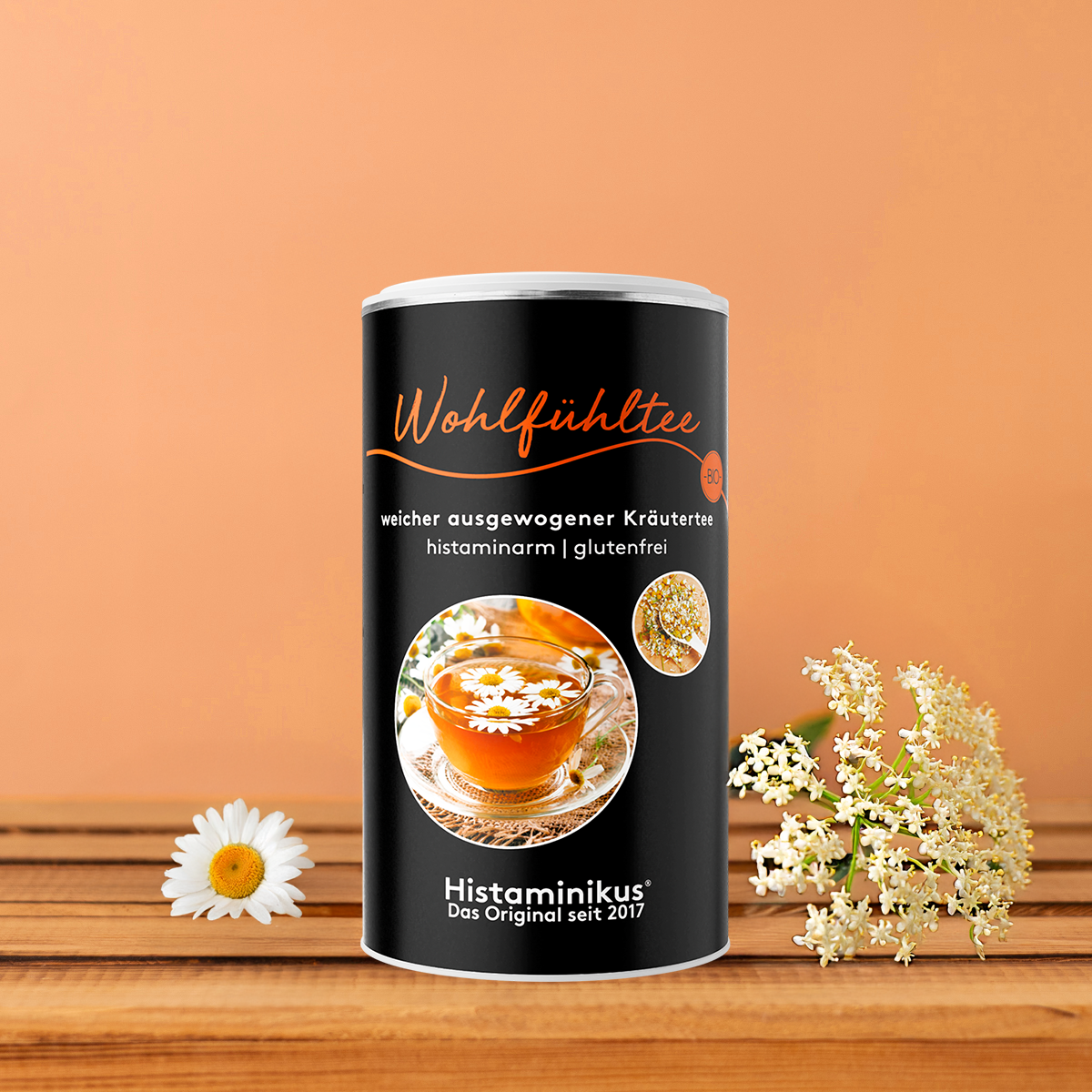 Schwarze Teedose „Wohlfühltee“ mit oranger Schrift und Abbildung einer Teetasse, dekoriert mit Holunderblüten, Kamille und Minze vor orangefarbenem Hintergrund. 