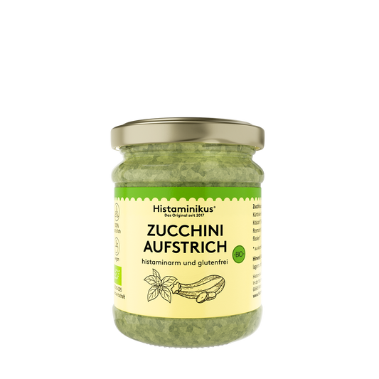 Glas mit goldfarbenem Deckel, gefüllt mit grünem Zucchini-Aufstrich. Auf dem cremefarbenen Etikett stehen Marke, Produktname und Hinweise wie „histaminarm und glutenfrei“, ergänzt durch ein kleines Bio-Siegel und eine gezeichnete Zucchini und Basilikum. Rechts und links sind weitere Produktinformationen angedeutet. 