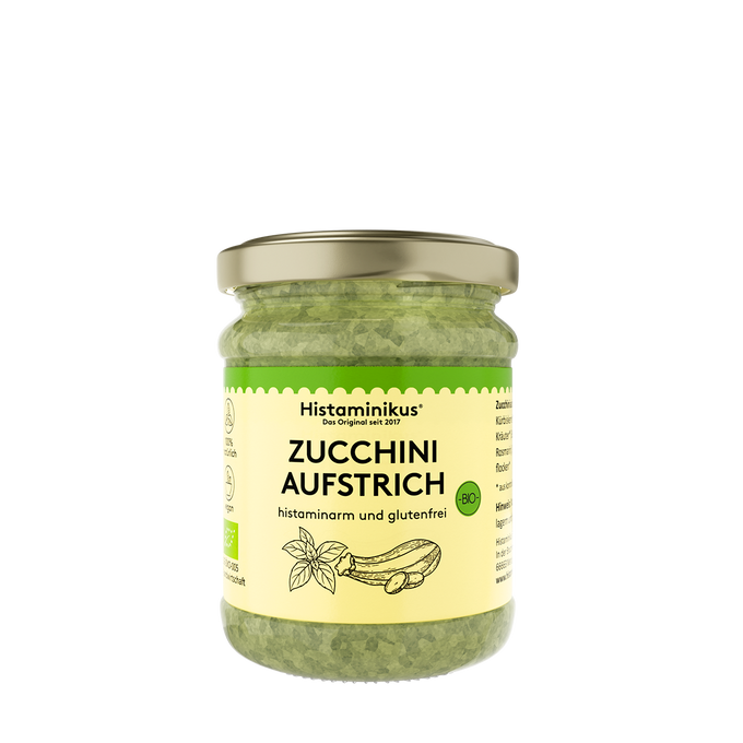 Glas mit goldfarbenem Deckel, gefüllt mit grünem Zucchini-Aufstrich. Auf dem cremefarbenen Etikett stehen Marke, Produktname und Hinweise wie „histaminarm und glutenfrei“, ergänzt durch ein kleines Bio-Siegel und eine gezeichnete Zucchini und Basilikum. Rechts und links sind weitere Produktinformationen angedeutet. 