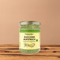 Glas Histaminikus Zucchini-Aufstrich vor einem warmen beigefarbenen Hintergrund auf einer Holzoberfläche. Das Glas steht frontal, das Etikett ist vollständig sichtbar.