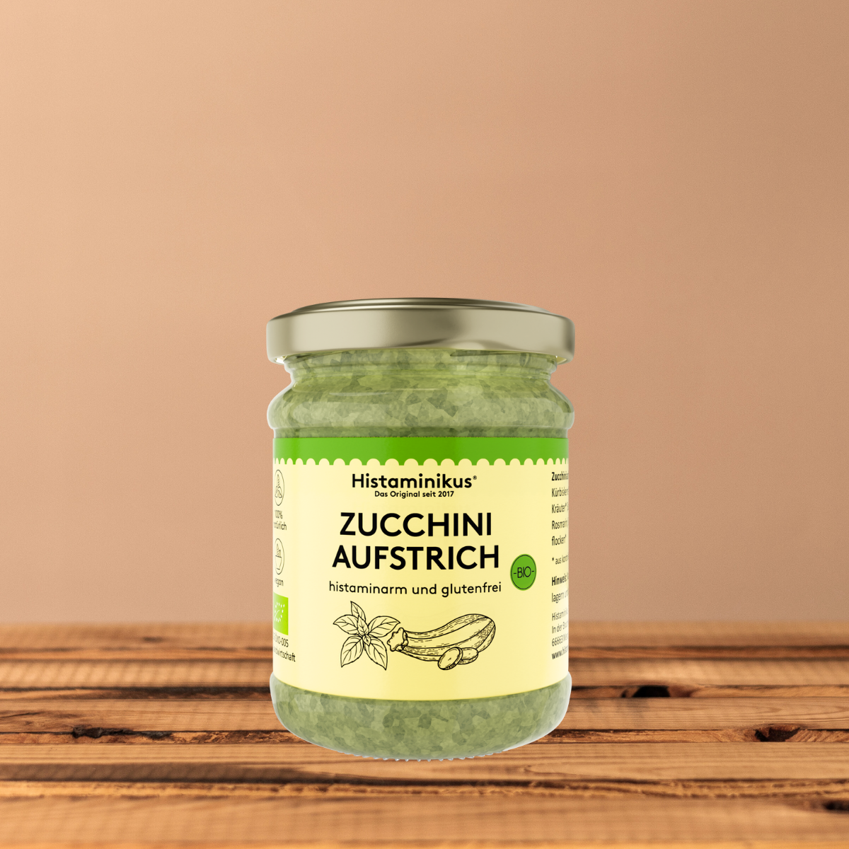 Glas Histaminikus Zucchini-Aufstrich vor einem warmen beigefarbenen Hintergrund auf einer Holzoberfläche. Das Glas steht frontal, das Etikett ist vollständig sichtbar.