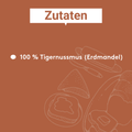 Grafik mit der Überschrift „Zutaten“ in einer weißen Sprechblase auf braunem Hintergrund. Darunter steht als einziger Stichpunkt „100 % Tigernussmus (Erdmandel)“. Im unteren rechten Bereich sind dezente lineare Illustrationen von Tigernüssen und aufgeschnittenen Erdmandeln im Hintergrund dargestellt.