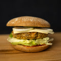 Burger mit runder Brötchenhälfte oben und unten. Zwischen den Brötchen ein pflanzlicher Patty mit sichtbaren Gemüseanteilen, darunter hellgrüner Blattsalat und rote Soße, darüber mehrere Scheiben roher Zwiebel. Der Burger steht auf einer Holzoberfläche, Hintergrund schwarz.