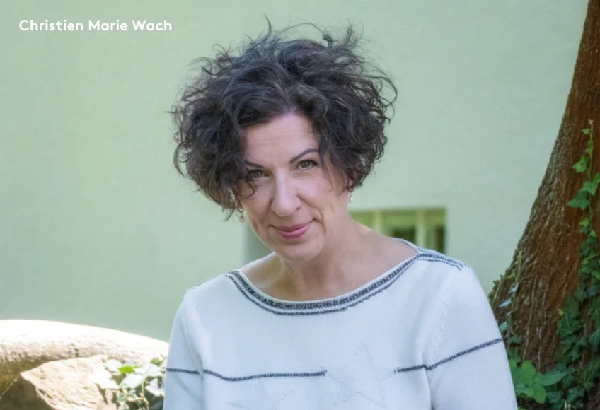 Porträtfoto von Christien Marie Wach mit lockigen dunklen Haaren, weißem Pullover mit dunklen Nähten, stehend neben einem Baum vor grünem Hintergrund.