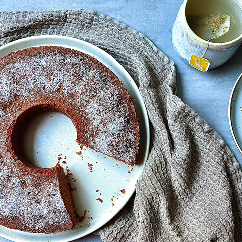 brauner Ringkuchen dekoriert mit einem grauen Tuch und einer Tasse Tee im Hintergrund