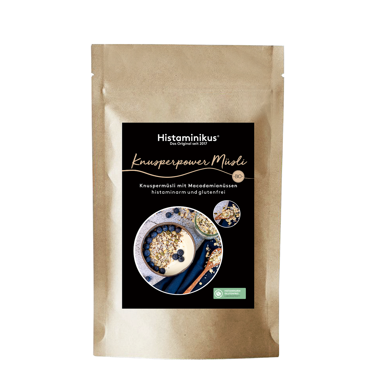 Produktfoto eines braunen Papierbeutels mit schwarzem Etikett. Darauf in weißer Schrift der Produktname „Knusperpower Müsli“ und die Beschreibung „Knuspermüsli mit Macadamianüssen – histaminarm und glutenfrei“. Abgebildet ist eine Müsli-Schale mit Joghurt und Blaubeeren.