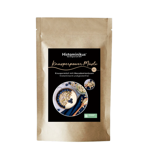Produktfoto eines braunen Papierbeutels mit schwarzem Etikett. Darauf in weißer Schrift der Produktname „Knusperpower Müsli“ und die Beschreibung „Knuspermüsli mit Macadamianüssen – histaminarm und glutenfrei“. Abgebildet ist eine Müsli-Schale mit Joghurt und Blaubeeren.