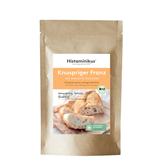 Produktabbildung der braunen Kraftpapierverpackung mit Etikett „Knuspriger Franz – Bio-Baguette-Mischung“. Untertitel: „histaminarm und glutenfrei – mit Hefe oder Backpulver backen“. Im unteren Bereich ein Foto von frisch gebackenem Baguette auf einem Holzbrett, mit dem handschriftlichen Zusatz „knusprig, kross, fluffig!“. Daneben grüne Siegel für „histaminarm glutenfrei“ und Bio-Qualität.