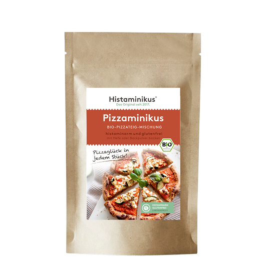 Brauner Standbodenbeutel mit zentralem, weiß-rot gestaltetem Etikett von Histaminikus; Produktname „Pizzaminikus – Bio-Pizzateig-Mischung“. Unterzeile „histaminarm und glutenfrei, mit Hefe oder Backpulver backen“. Rechts am Etikett ein grünes BIO-Siegel. Darunter Produktfoto: runde Pizza, in Stücke geschnitten, mit weißem Belag und Basilikum; daneben handschriftlicher Spruch „Pizzaglück in jedem Stück!“. Unten rechts ein grünes Siegel „Histaminarm – Glutenfrei – Laborgeprüft“.
