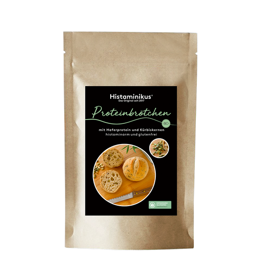 Vorderansicht einer braunen, stehenden Papiertüte mit Produktetikett „Histaminikus Proteinbrötchen“. Auf dem Etikett sind zwei runde, aufgeschnittene Brötchen mit sichtbaren Kürbiskernen sowie ein Brotmesser auf einem Holzbrett abgebildet. Die Beschriftung auf dem Etikett lautet: „mit Haferprotein und Kürbiskernen, histaminarm und glutenfrei“. Rechts oben auf dem Etikett ist ein kleines rundes Foto mit Kürbiskernen und Teigstücken zu sehen.