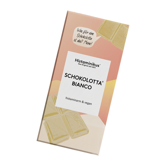 Schräg dargestellte Verpackung der veganen, histaminarmen Schokolade „Schokolotta Bianco“ von Histaminikus mit weißem Etikett, handschriftlicher Sprechblase, weißen Schokoladenstücken.