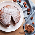 Runder Kuchen mit Puderzucker bestäubt auf weißem Teller, ein Stück herausgeschnitten. Daneben braune Haselnüsse auf blauer Stoffserviette.