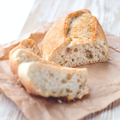Nahaufnahme eines frisch gebackenen Baguettes auf braunem Papier: Im Vordergrund zwei angeschnittene Scheiben mit luftiger Krume, dahinter ein goldbraun gebackenes Baguettestück mit knuspriger Kruste. Die helle Holzoberfläche und das weiche Licht unterstreichen den natürlichen, handgemachten Charakter.