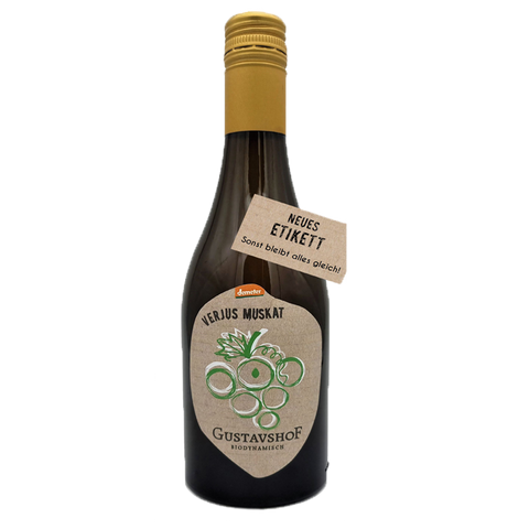 Braune Glasflasche mit goldfarbenem Schraubverschluss. Auf dem Etikett steht „VERJUS MUSKAT“ sowie „Gustavshof – Biodynamisch“ mit einer stilisierten grünen Traubengrafik. Ein zusätzlicher Anhänger am Flaschenhals trägt die Aufschrift „NEUES ETIKETT – Sonst bleibt alles gleich!“.