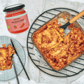 Rechts eine Auflaufform mit Lasagne auf einem schwarzen Kuchengitter. Am linken Bildrand ein schwarz-weißer Teller mit Lasagne. In der oberen linken Bildecke ist ein Glas der No!Mato Sosse von Histaminikus zu sehen.